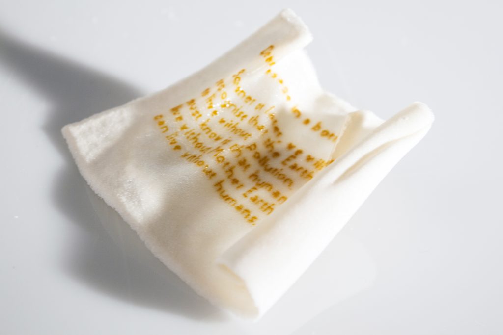 Art piece "Unnamed" in 2018 by Alise Stopina. Glass blown prefume bottles with cork stopper and white glass powder paperlike pieces with hand written messeages on them, to simulate "message in a bottle". Zoomed in one specific message.
