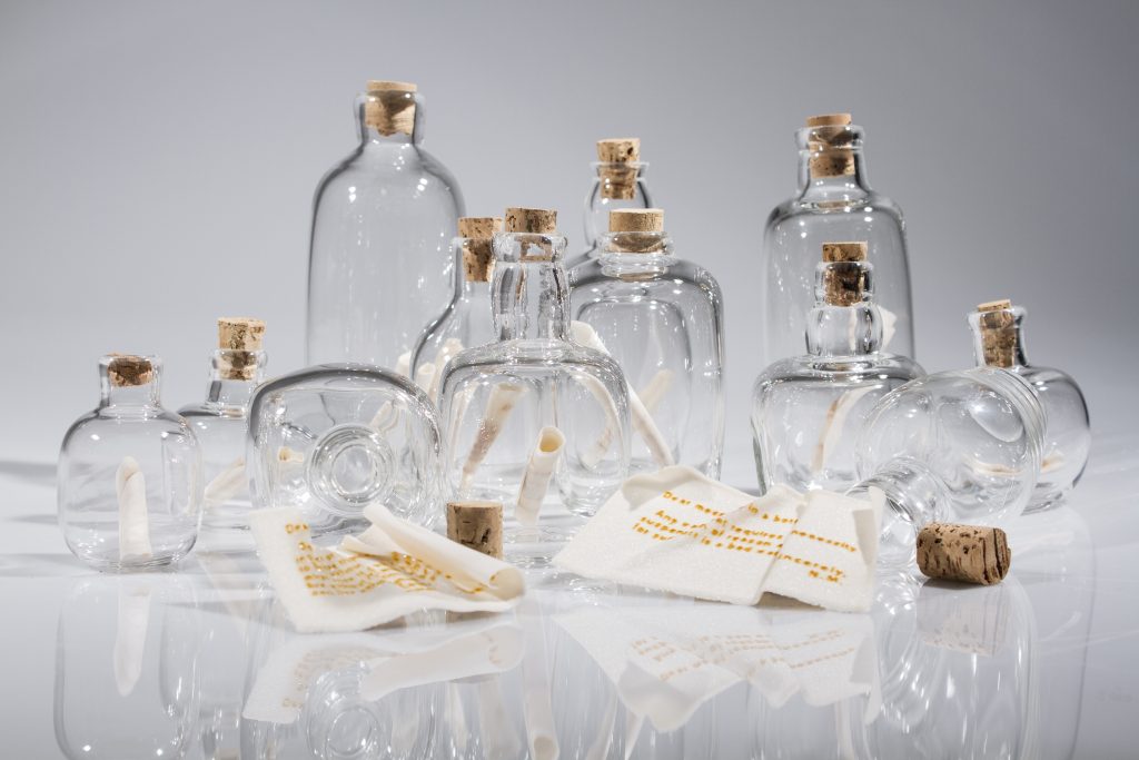 Art piece "Unnamed" in 2018 by Alise Stopina. Glass blown prefume bottles with cork stopper and white glass powder paperlike pieces with hand written messeages on them. Collection of all together.