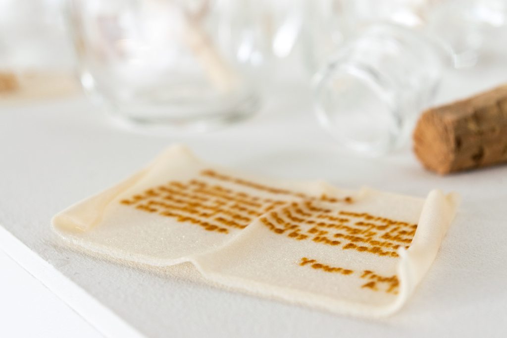 Art piece "Unnamed" in 2018 by Alise Stopina. Glass blown prefume bottles with cork stopper and white glass powder paperlike pieces with hand written messeages on them, to simulate "message in a bottle". Zoomed in one specific message.